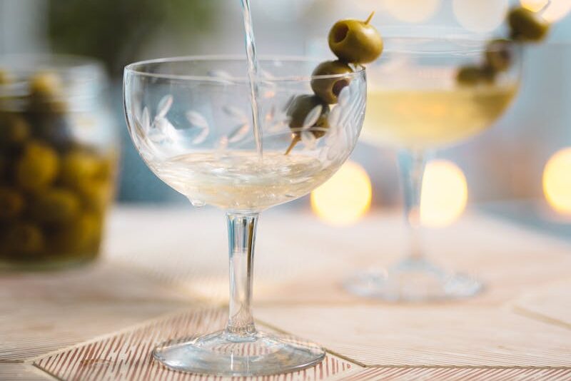 A classic vodka martini being poured from a shaker into a glass garnished with green olives.