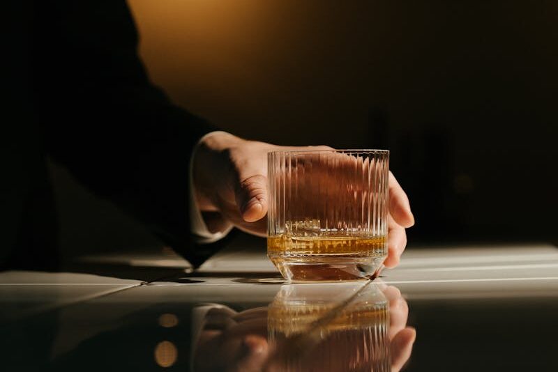 Close-up of a hand holding a whiskey glass with reflections on a bar counter in a dim light.