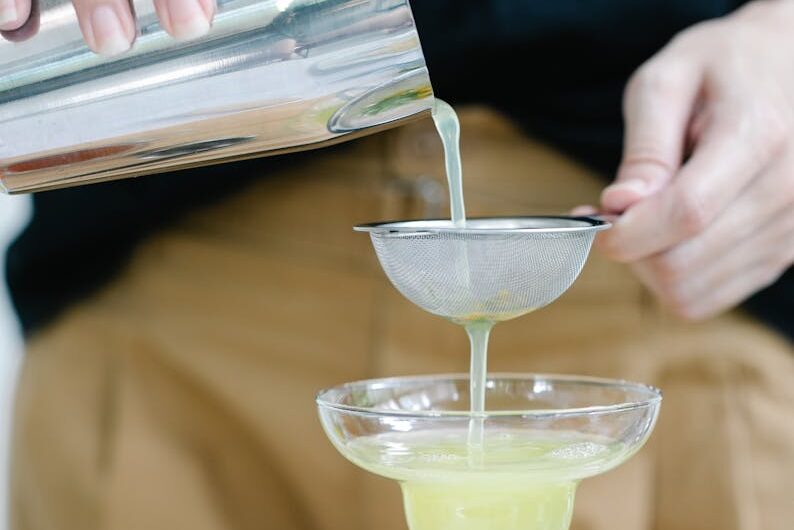 A person pouring a freshly shaken cocktail through a sieve into a glass on a wooden table.