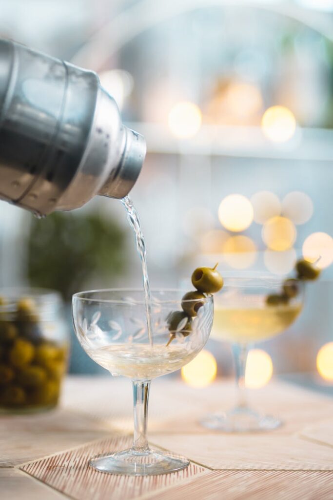 A classic vodka martini being poured from a shaker into a glass garnished with green olives.